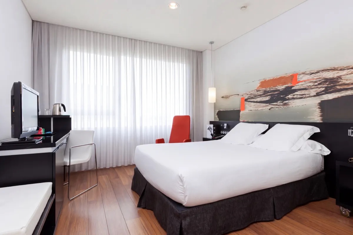 Hotel room with a white-made double bed, black bed skirt, an orange armchair, sheer curtains over a large window and a wood-effect floor.