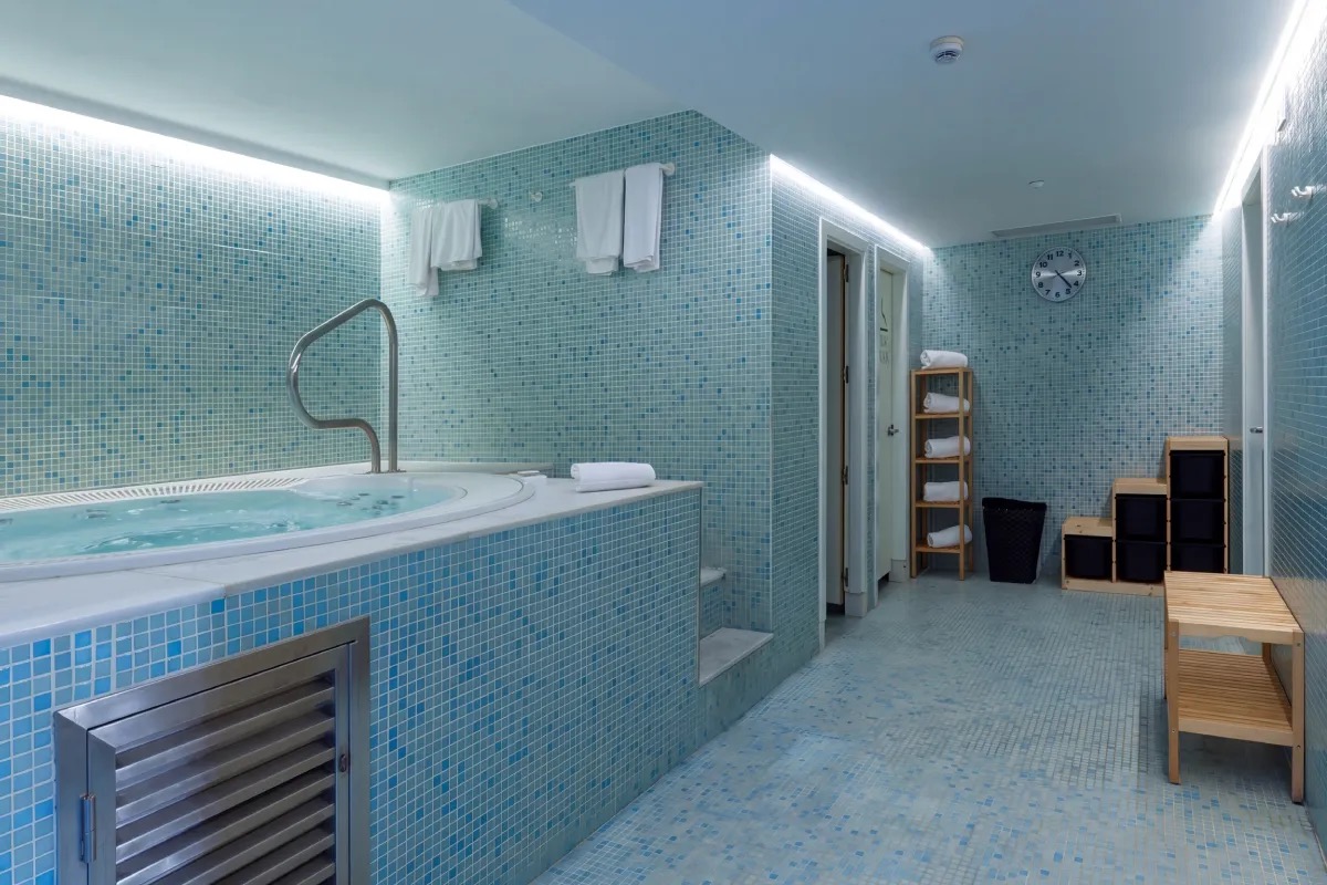 Blue-tiled wellness area with a round jacuzzi tub, a handrail, folded towels and a wall clock.