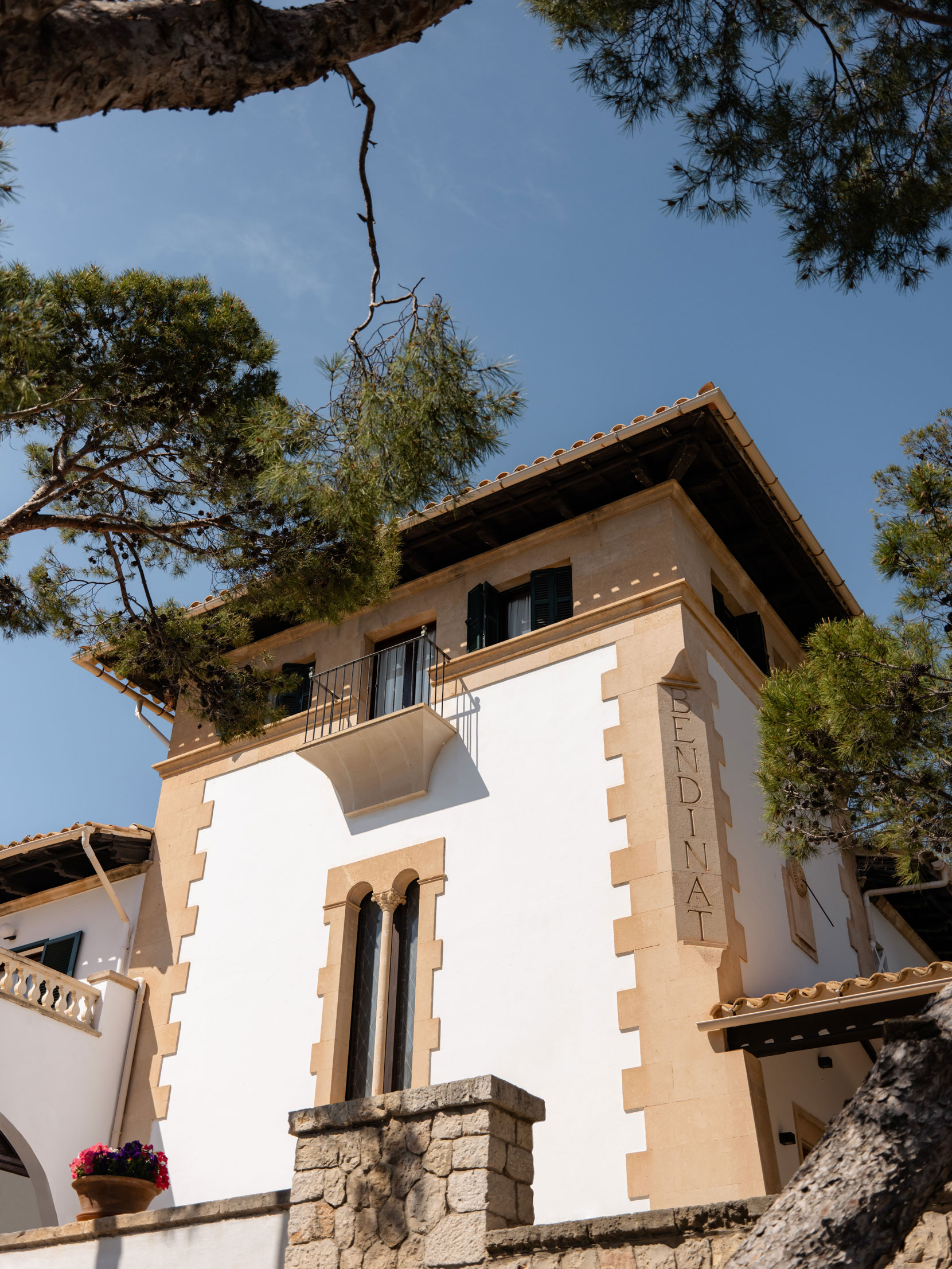Upward view of the stone-and-white facade carved with the BENDINAT name, framed by pine trees