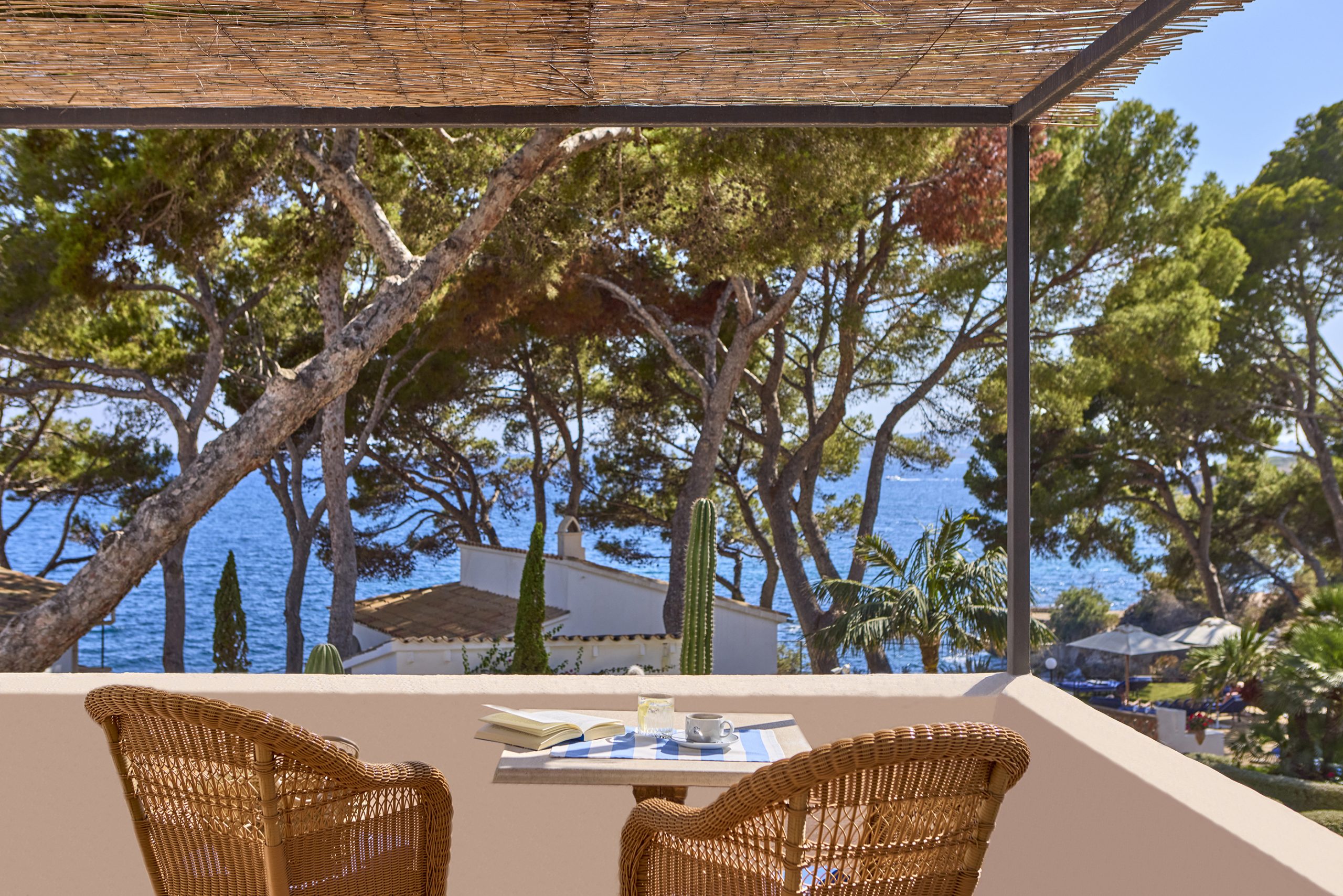Pergola-shaded terrace table set for coffee and reading, with pine trees and the sea beyond