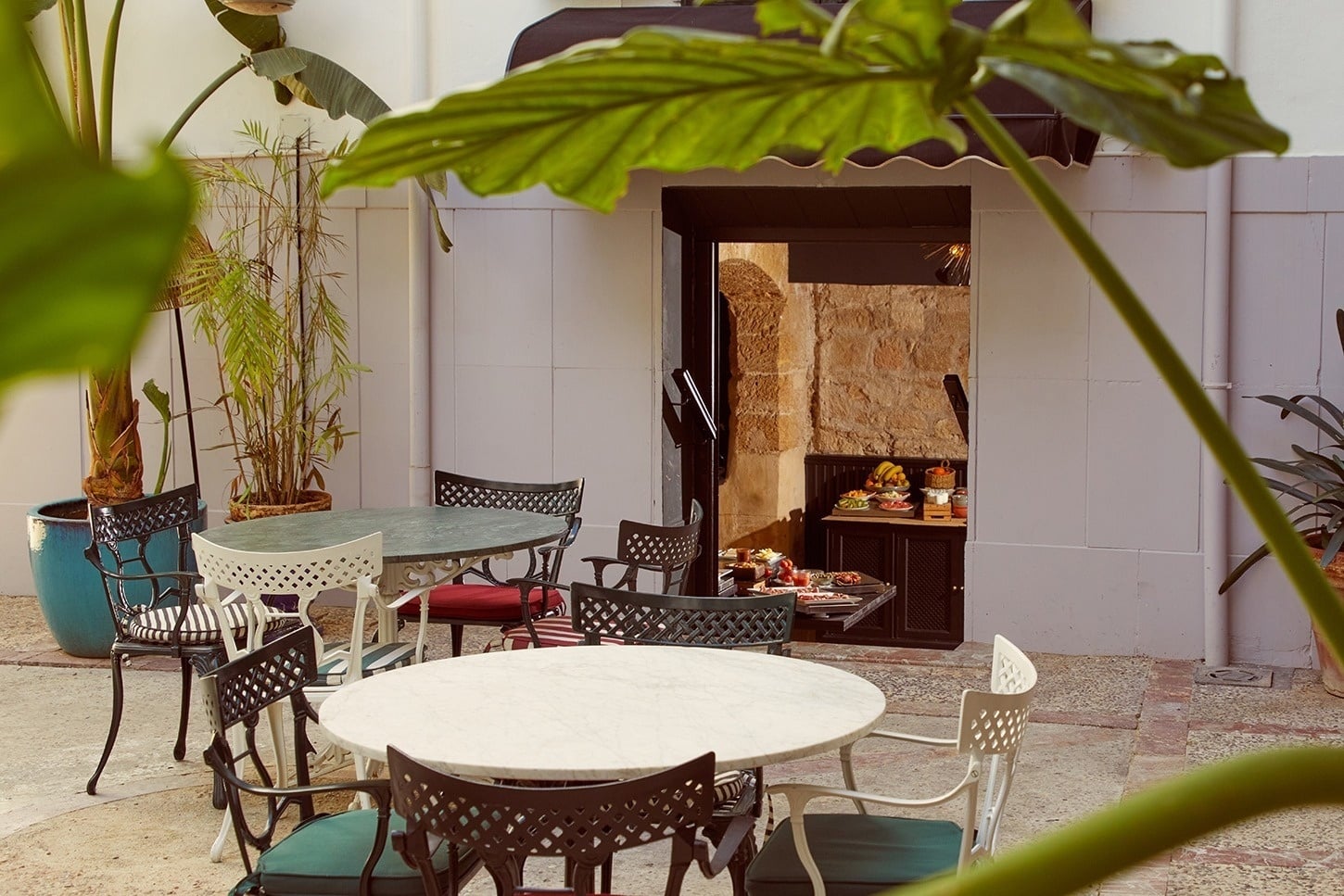 Courtyard breakfast nook with a stone alcove set up as a buffet station, seen past leafy plants and outdoor tables
