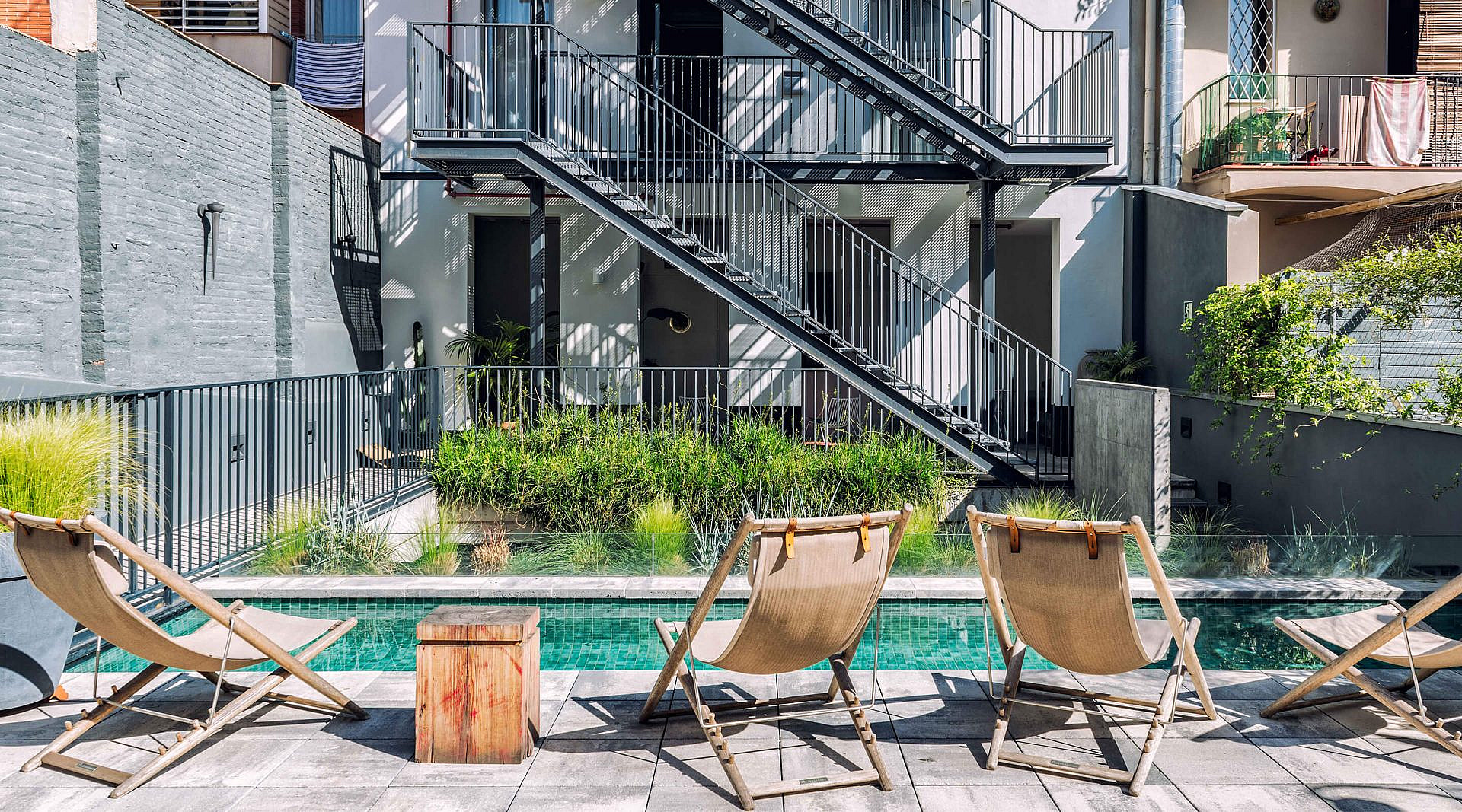 Wooden deck chairs on the paving beside the courtyard pool, with a metal staircase and grasses planted behind