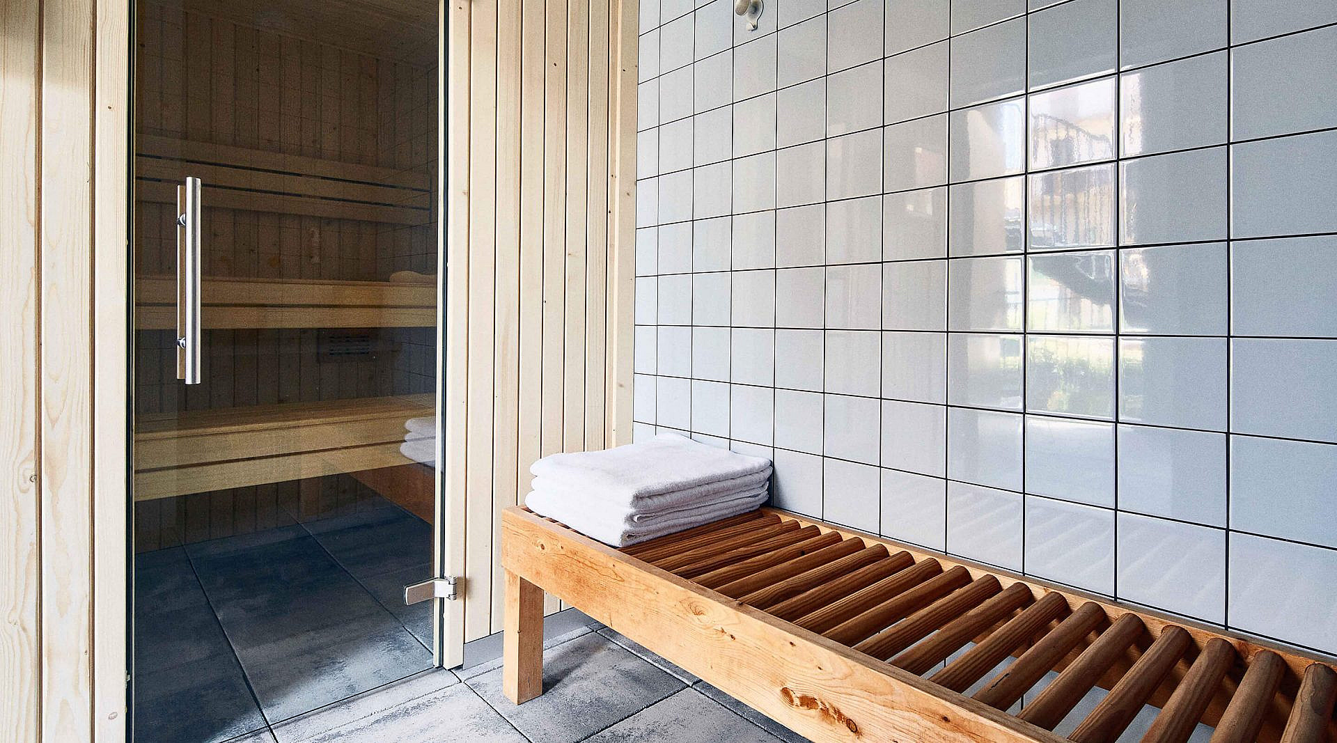 Dry sauna behind a glass door, with a slatted wooden bench and folded towels against white tiling