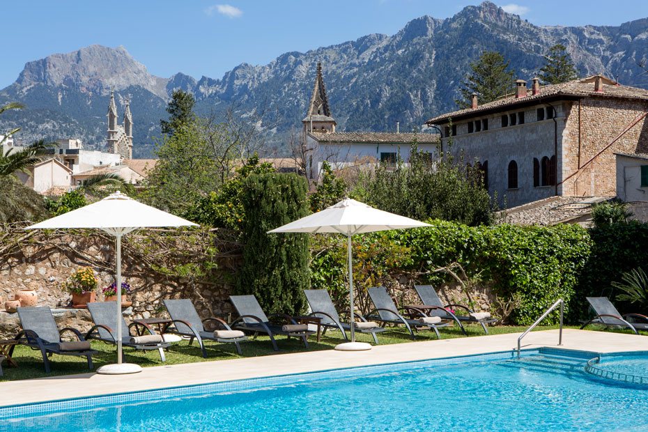 Outdoor pool with sun loungers and umbrellas, backed by a stone wall, church tower and the Serra de Tramuntana.