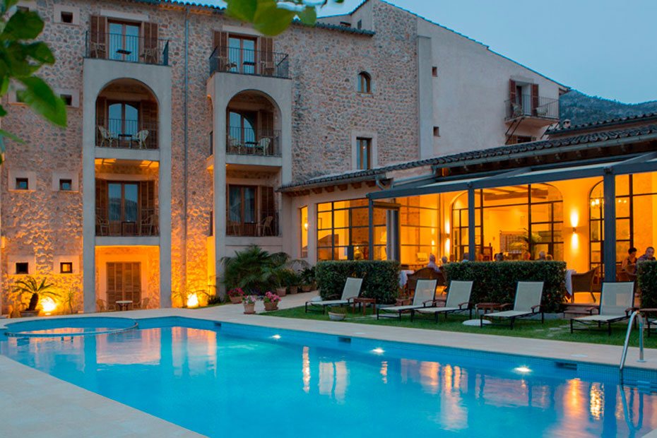 Evening view of the hotel's stone facade and lit dining terrace beside the illuminated pool.