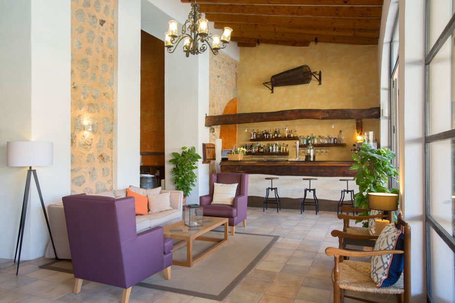 Hotel bar and lounge with a wood-beamed ceiling, chandelier, purple armchairs and a stone wall.