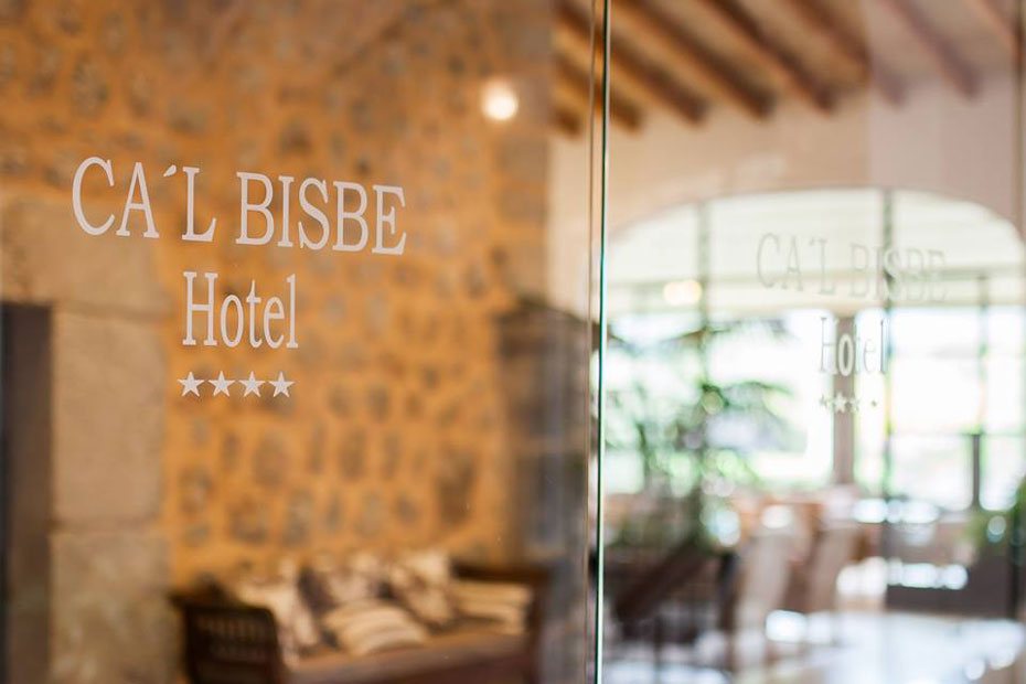 Glass entrance doors etched with the Ca'l Bisbe Hotel name, opening onto the lobby seating area.