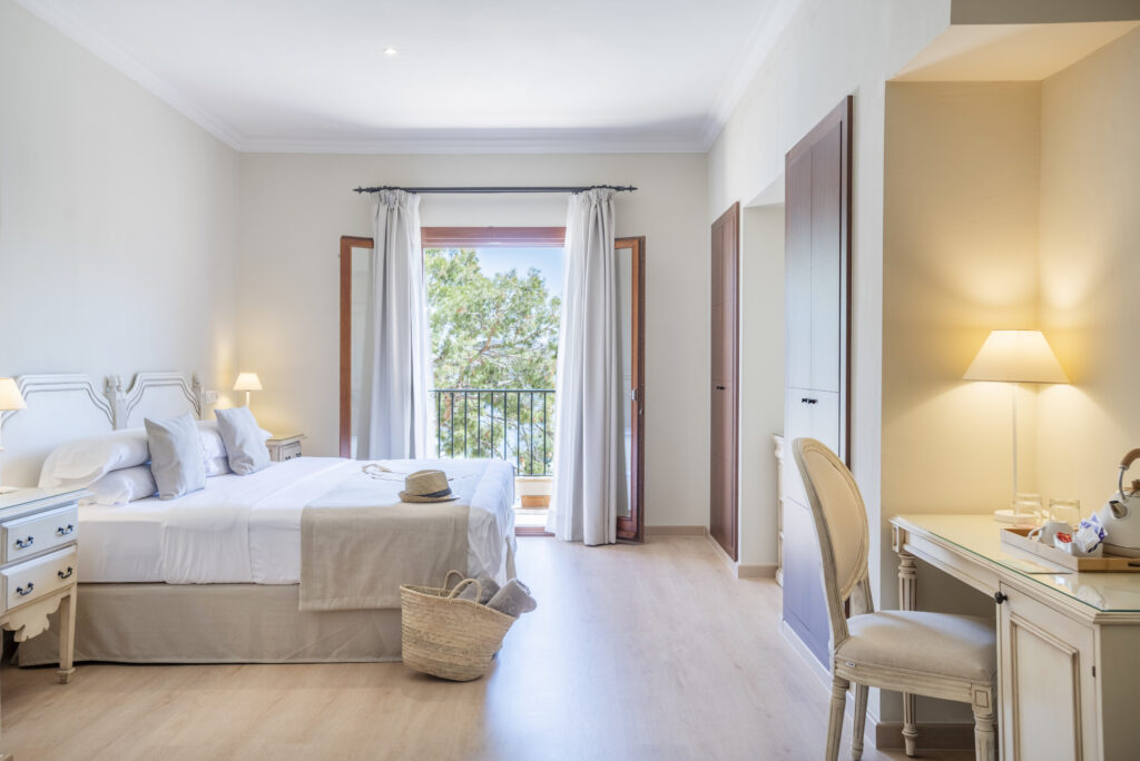 Bedroom at Hotel Cala Fornells with a white-framed bed, cream linens and open balcony doors framed by curtains.