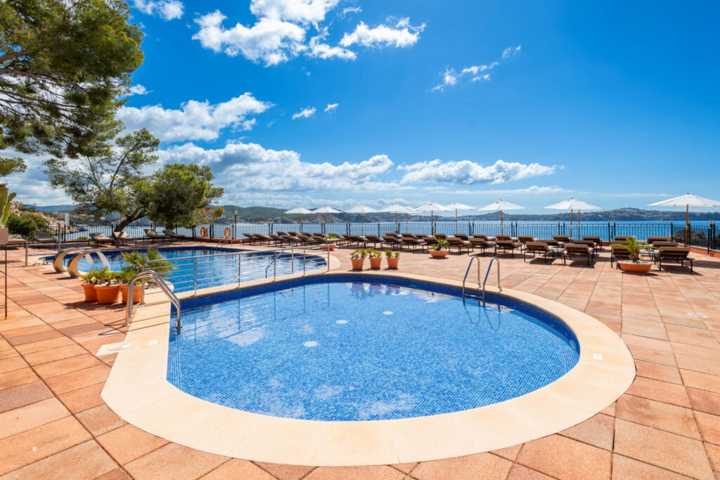 Outdoor pool terrace at Hotel Cala Fornells with sun loungers, white umbrellas and a view over the bay.