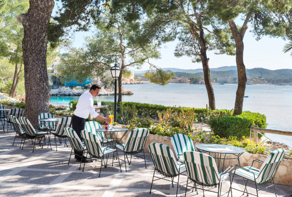 Seaside terrace at Hotel Cala Fornells with striped chairs under pine trees and a waiter setting down a drink, the cove visible behind.