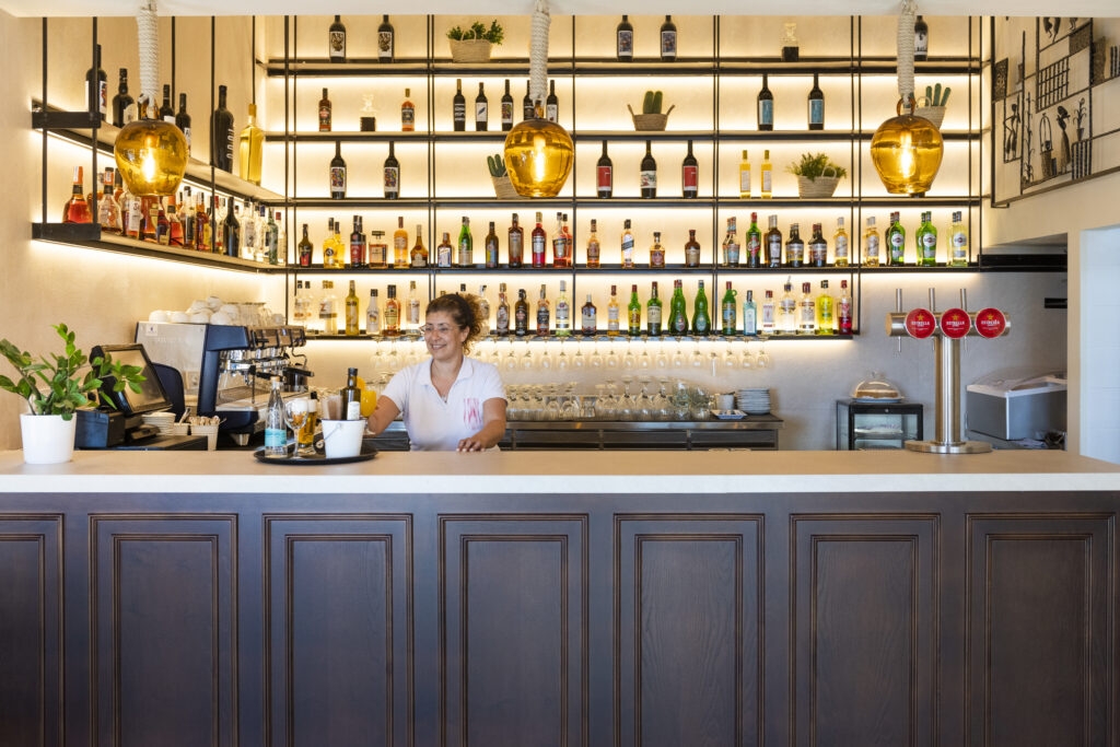 Hotel bar at Hotel Cala Fornells with backlit shelves of bottles and a staff member behind the wooden counter.