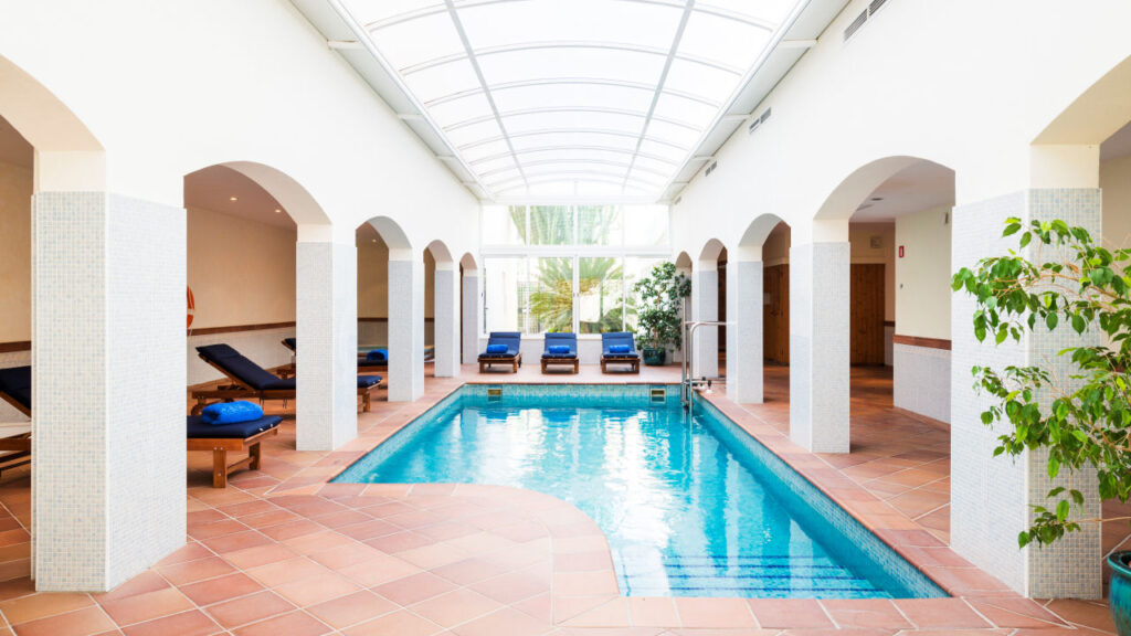 Indoor wellness pool at Hotel Cala Fornells under a skylight, framed by stone archways and loungers.
