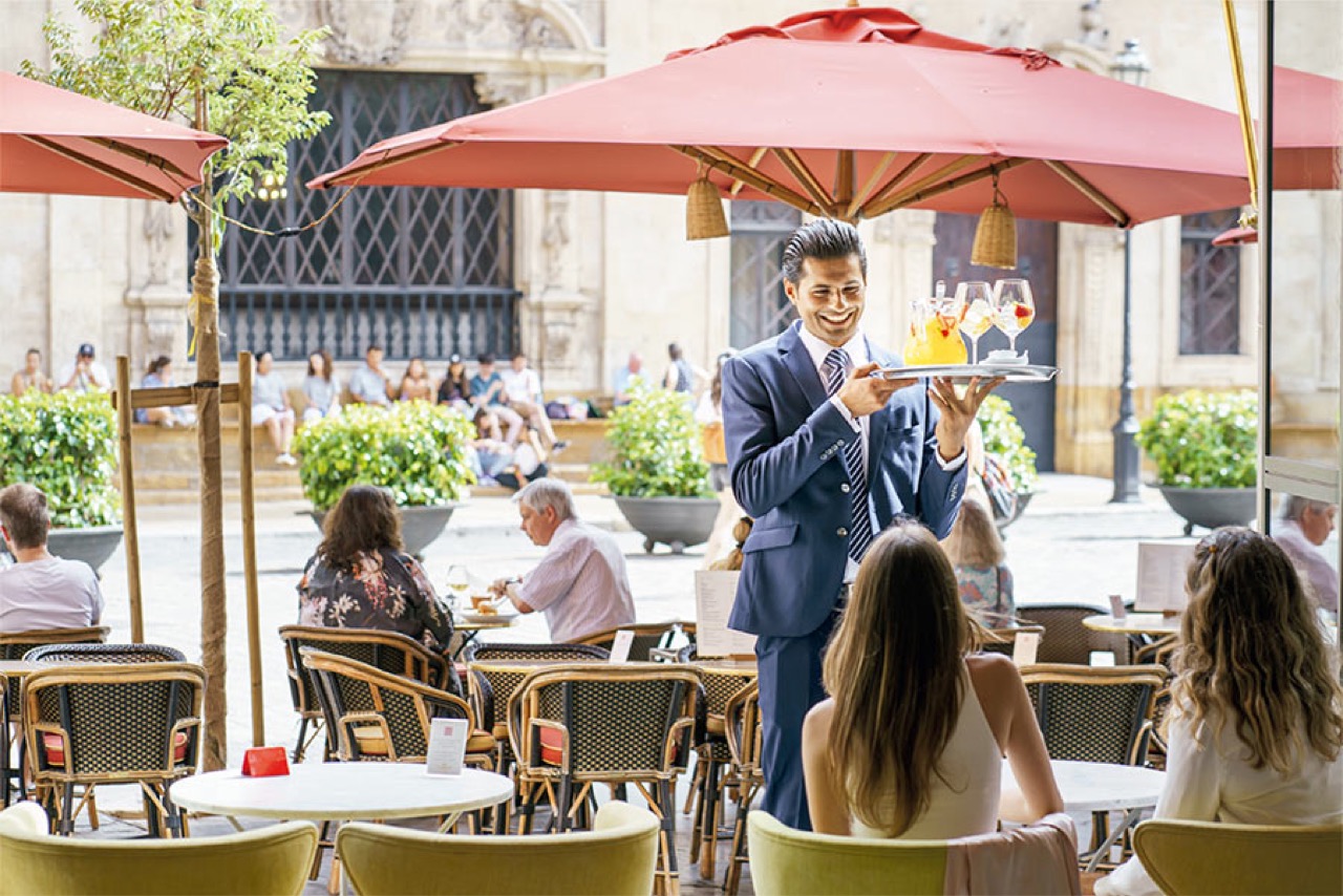 Outdoor terrace seating under red umbrellas facing the plaza