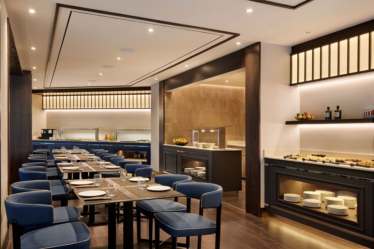 A buffet breakfast room with blue upholstered chairs at long tables, facing a dark-blue buffet counter and a show-cooking station.