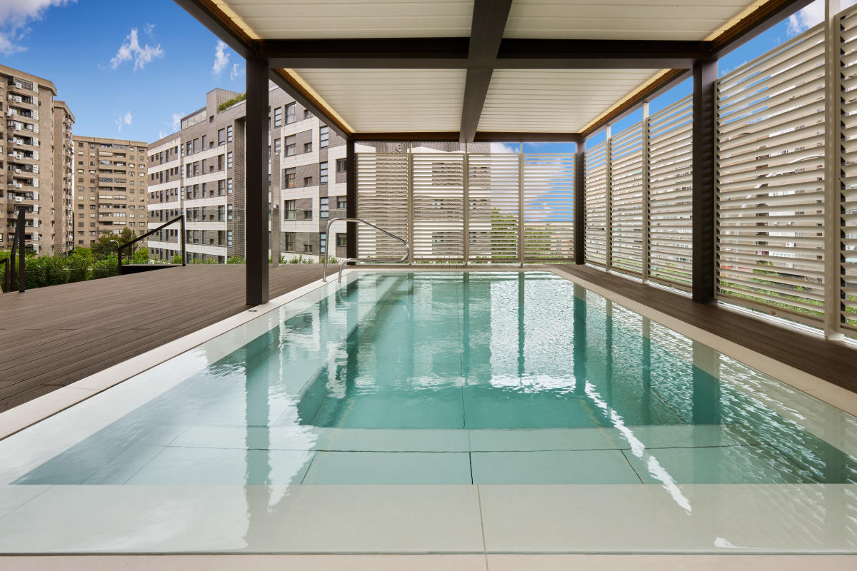 A covered rooftop pool with a wood-decked surround and slatted privacy screens, overlooking nearby apartment buildings.