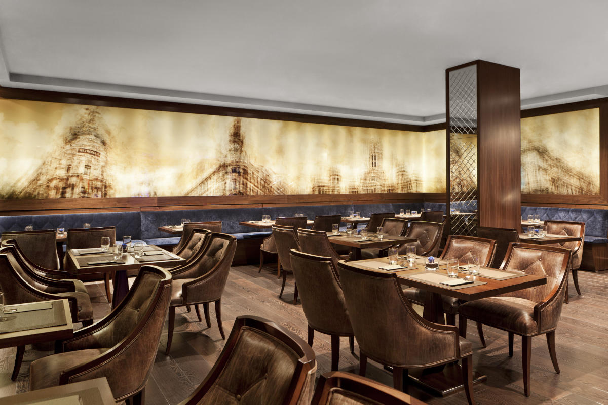 The Claridge Café Restaurant dining room, with leather chairs at set tables in front of a long sepia-toned mural of Madrid landmarks.