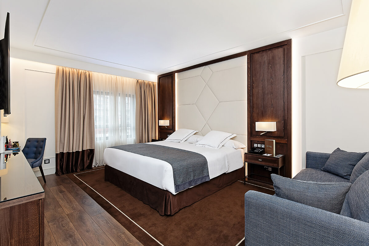 A hotel room with a wide bed against a padded cream headboard, wood panelling, a grey sofa, and a desk with a mirror.