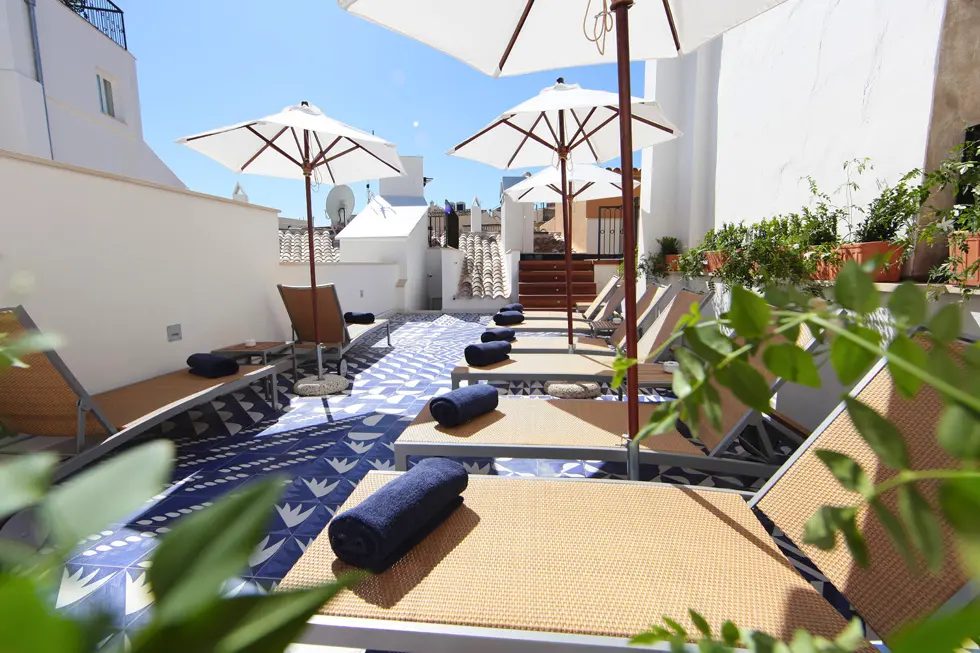 Rooftop lounging terrace at Hotel Cort with white sun umbrellas, blue-patterned mats, and loungers overlooking Palma's rooftops