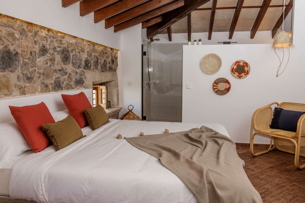 Bedroom with an exposed stone accent wall, white bedding with red and olive cushions, wood-beamed ceiling and an en-suite glass shower.