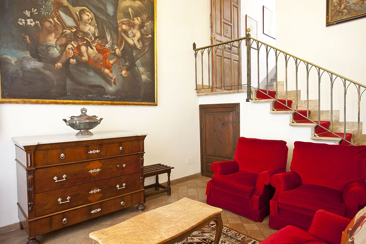 Sitting room at Hotel Dalt Murada with red velvet armchairs, an antique marble-topped chest and a brass-railed stone staircase