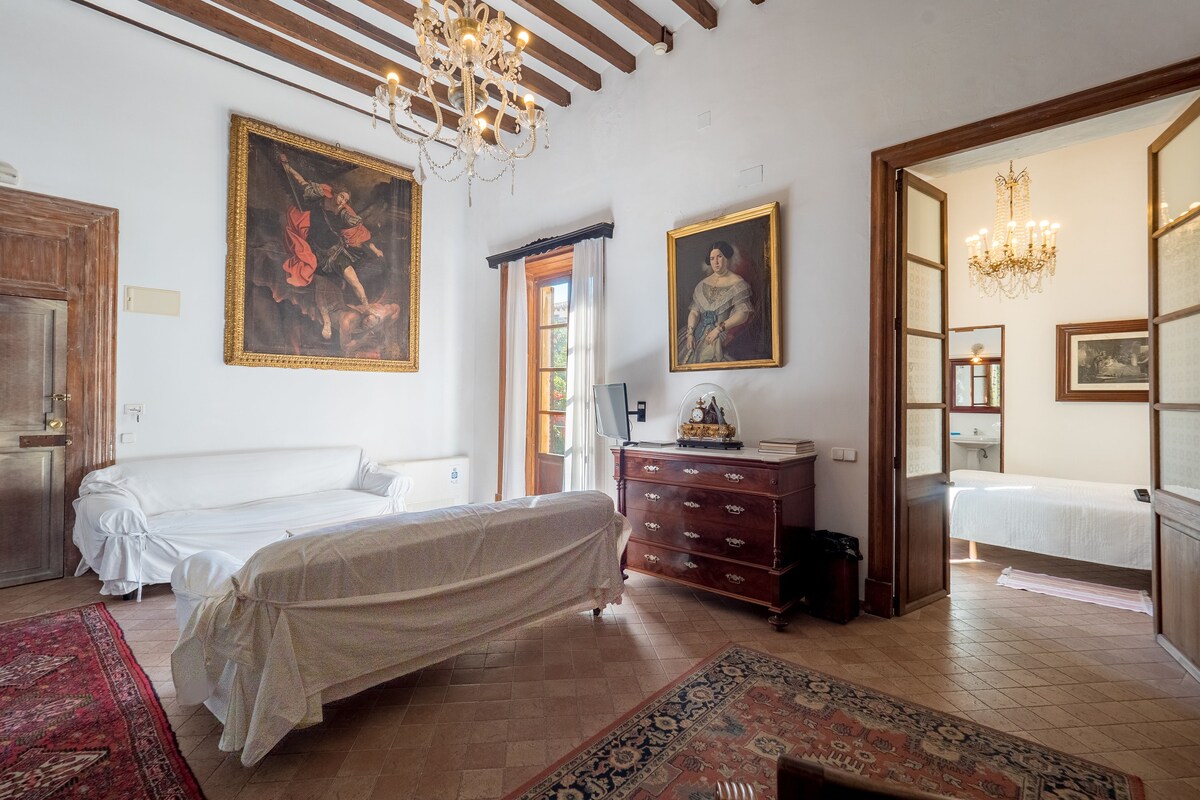 Spacious guest room at Hotel Dalt Murada with a glass chandelier, exposed wooden beams, terracotta floors and antique oil paintings