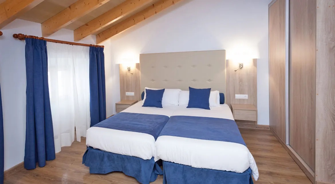 A twin room at Hotel El Guía with a sloped wood-beamed ceiling, a beige upholstered headboard and blue bedspreads.