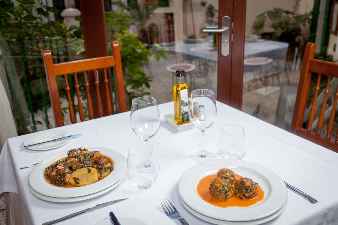 A table at Ca's Pentinadó set with two Mallorcan dishes, olive oil and wine glasses, beside glass doors onto the hotel courtyard.