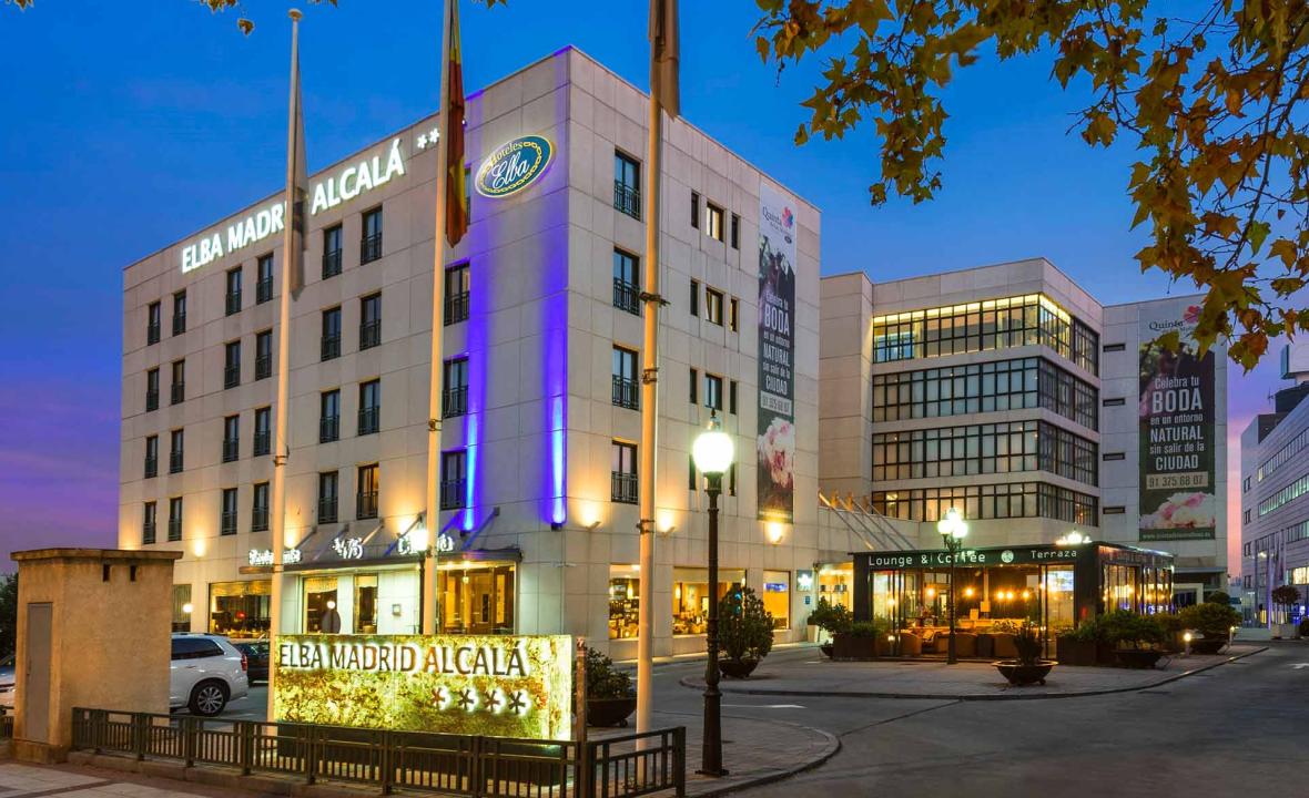 The Elba Madrid Alcalá building lit up at dusk, with its illuminated sign, flagpoles, and a ground-floor lounge and coffee terrace.