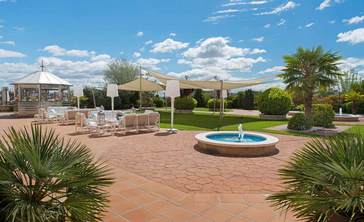 An outdoor garden terrace with a small round fountain, white lounge sofas under a shade sail, potted palms, and a gazebo bar to the left.
