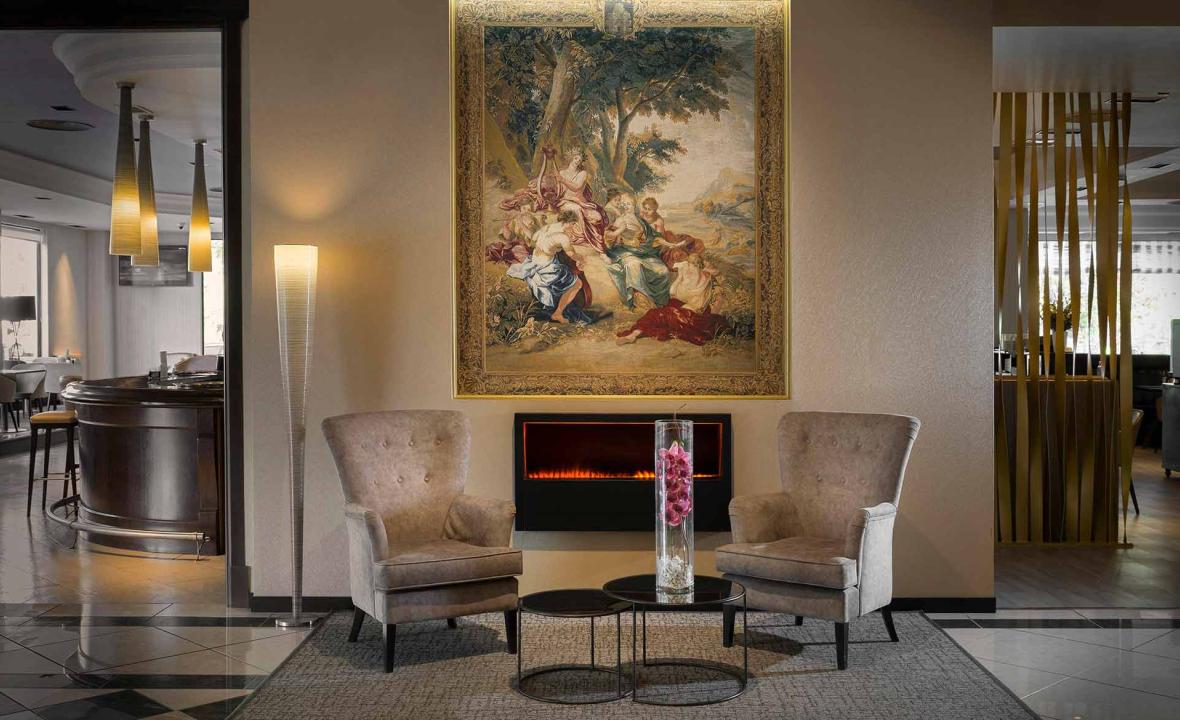 A lobby lounge with two beige armchairs facing an electric fireplace under a large framed tapestry, with the hotel bar visible to the left.