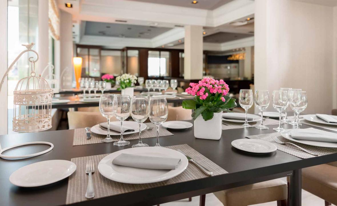 A restaurant dining room with tables set in white linens, wine glasses, and small vases of pink flowers, with a decorative birdcage ornament in the foreground.