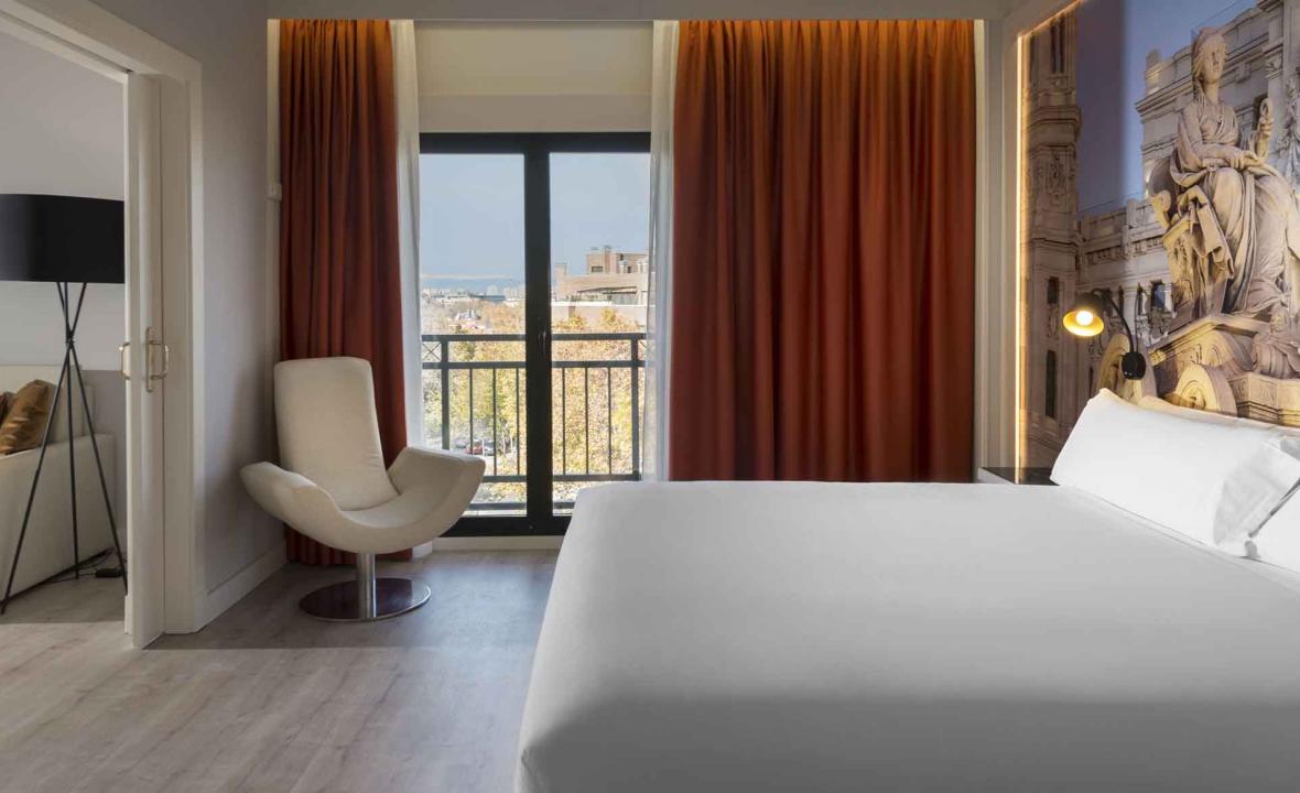 A hotel room with a white bed against a wall mural of a Madrid monument, an orange curtain, a cream armchair, and a balcony with a city view.