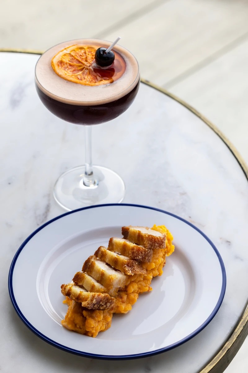 A cocktail garnished with a dried orange slice and cherry next to a plate of sliced pork belly on mashed pumpkin, on a marble table.