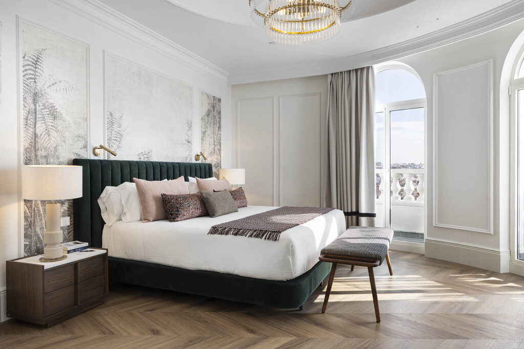A hotel room with a dark green upholstered headboard, arched window and small balcony overlooking Madrid rooftops.