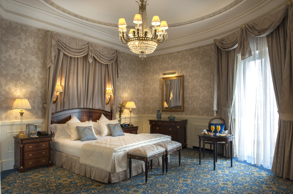 A classic suite with a canopied bed, crystal chandelier, patterned blue carpet and a small breakfast table by the window.