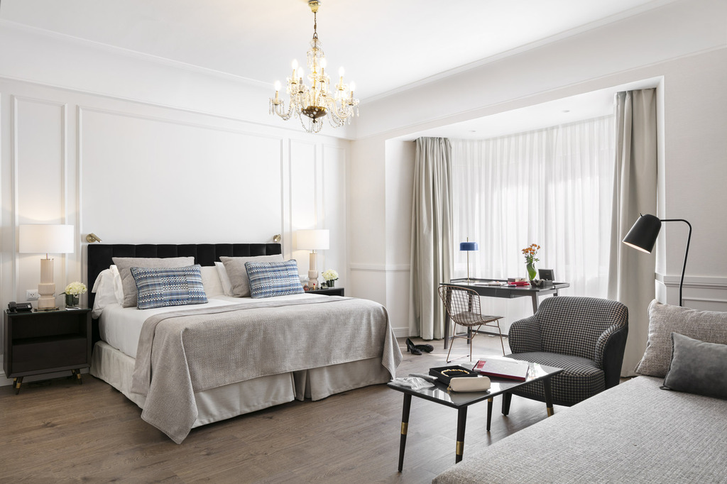 A modern suite with a dark tufted headboard, a seating area with an armchair and sofa, and a bay window nook with a desk.