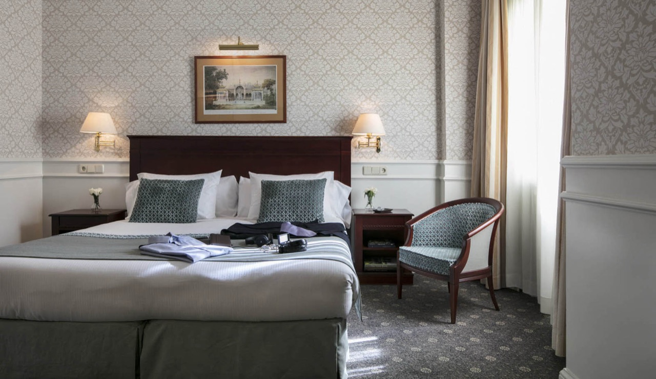 A classic hotel room with damask wallpaper, a dark wood headboard, a framed print, and a patterned armchair by the curtained window.
