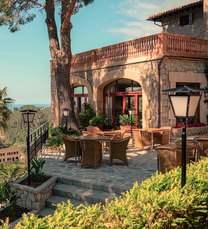 Restaurant terrace framed by stone arches, with wicker chairs set beneath a pine tree and a hillside view