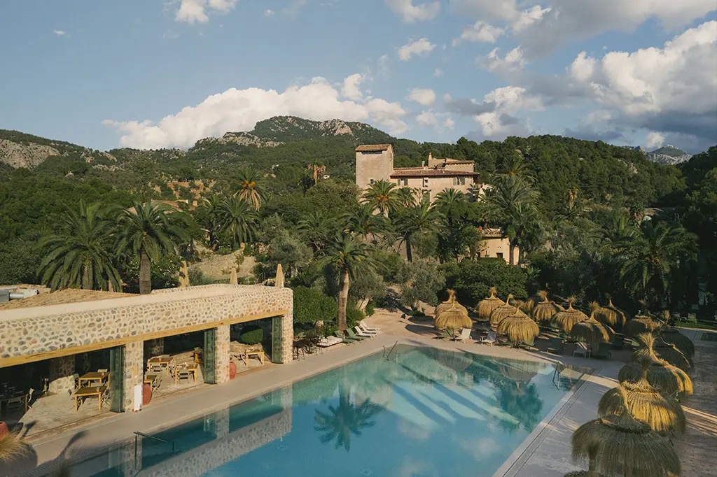 Aerial view of the outdoor pool terrace with straw umbrellas, the stone finca building and the Tramuntana mountains behind