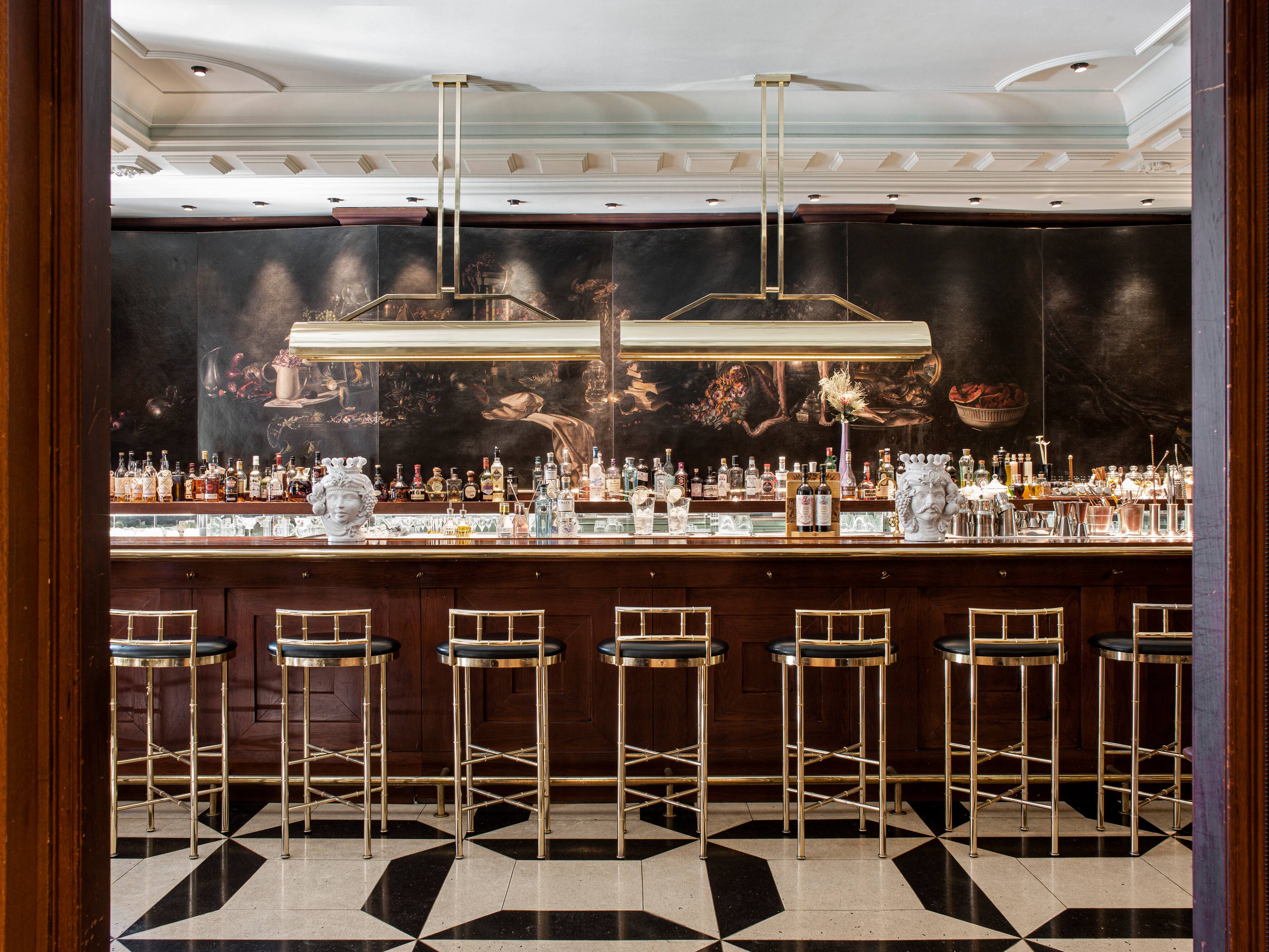 A bar with a dark wood counter, brass bamboo-style stools with black seats, a backlit row of spirits bottles beneath a dark still-life mural and a black-and-white geometric tiled floor.