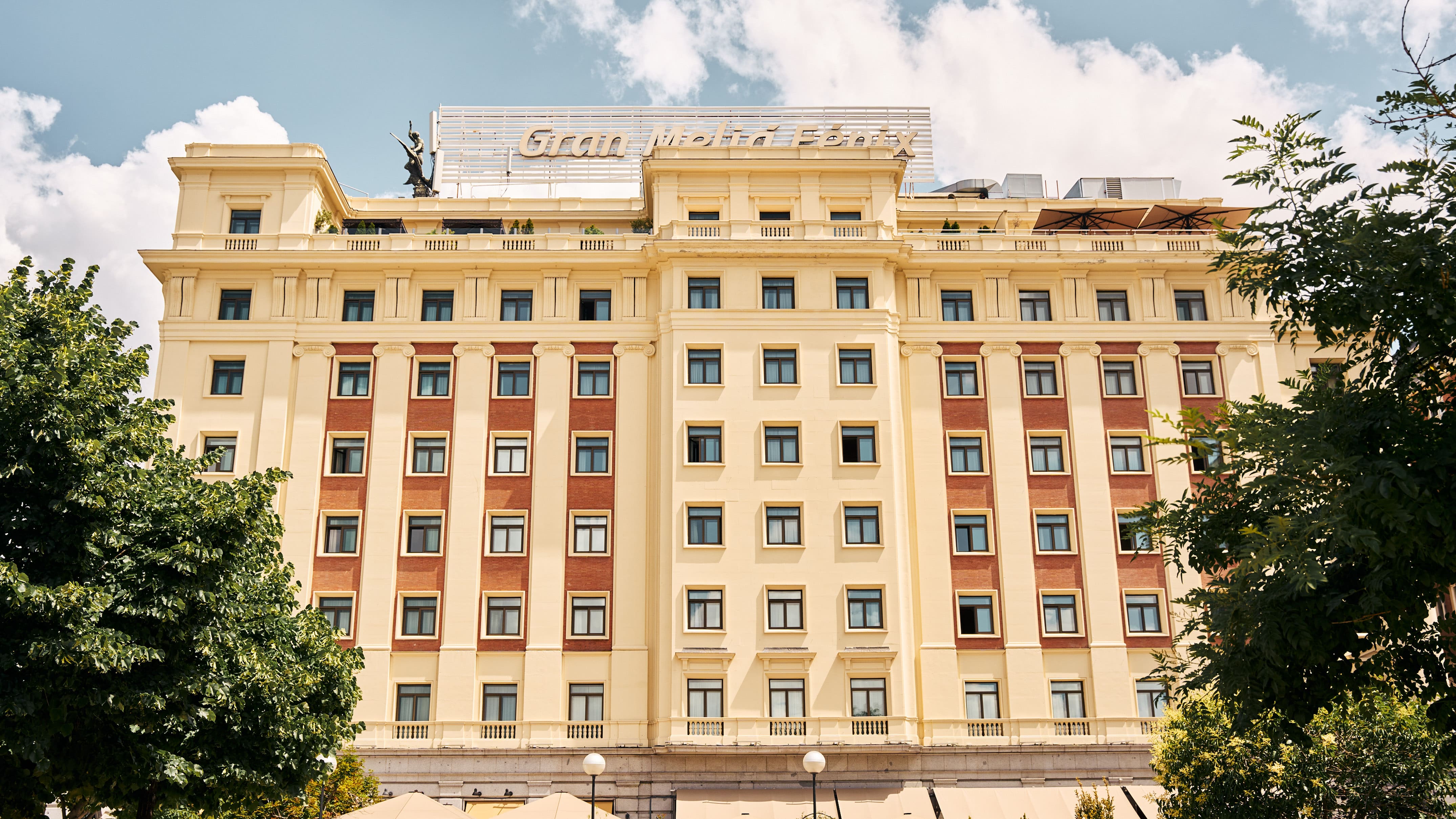 A cream and red-brick hotel facade of several storeys with balustraded balconies, a rooftop sign reading Gran Melia Fenix and a winged statue on the roofline, framed by green trees.