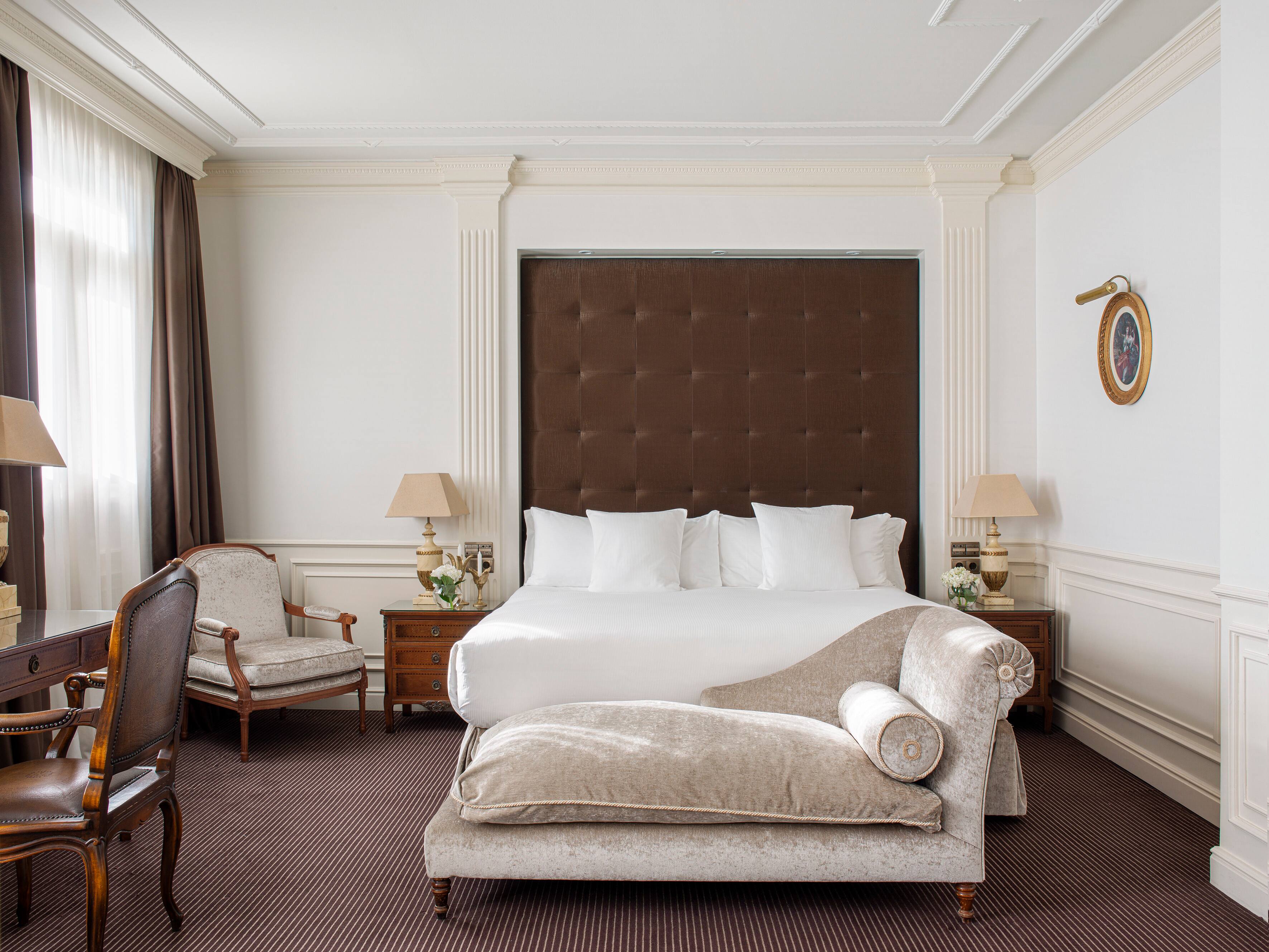 A guest room with a tall brown tufted headboard, white bedding, twin shaded lamps on inlaid wooden nightstands, a velvet chaise longue at the foot of the bed and striped brown carpet.
