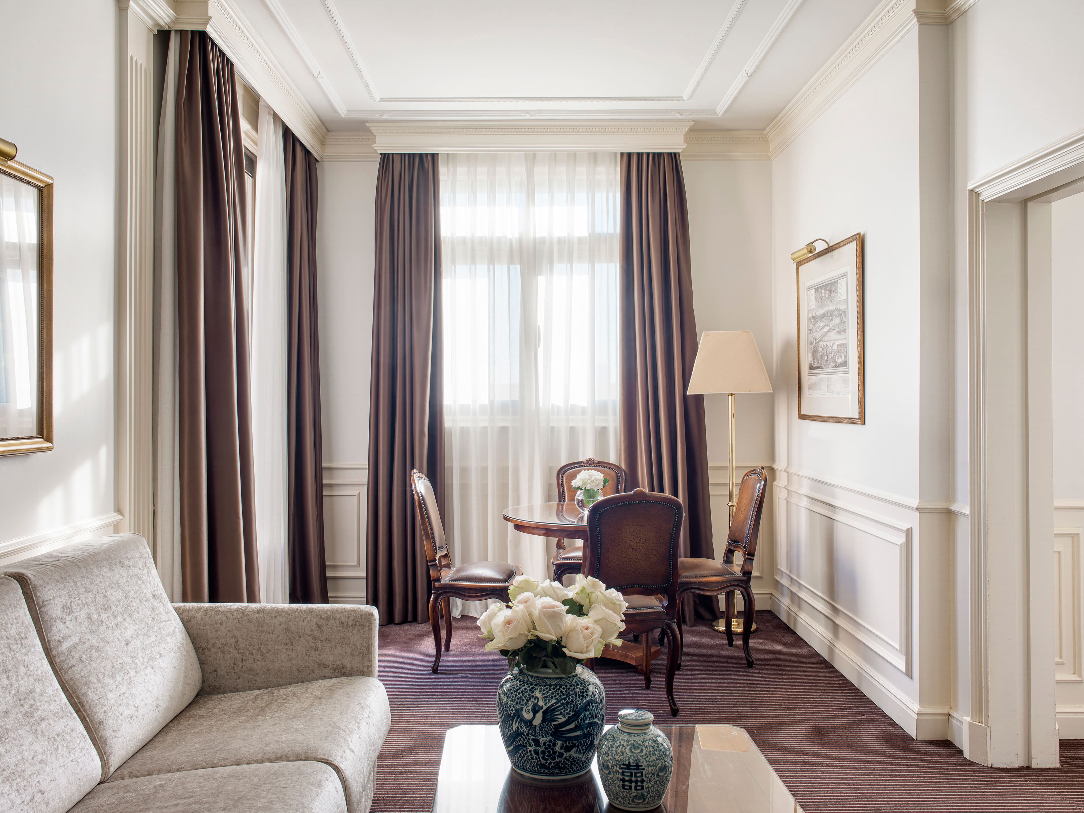 A sitting room with white wainscoting and moulded cornicing, floor-length brown curtains, a pale velvet sofa, a round wooden table with four antique-style chairs and a brass floor lamp.