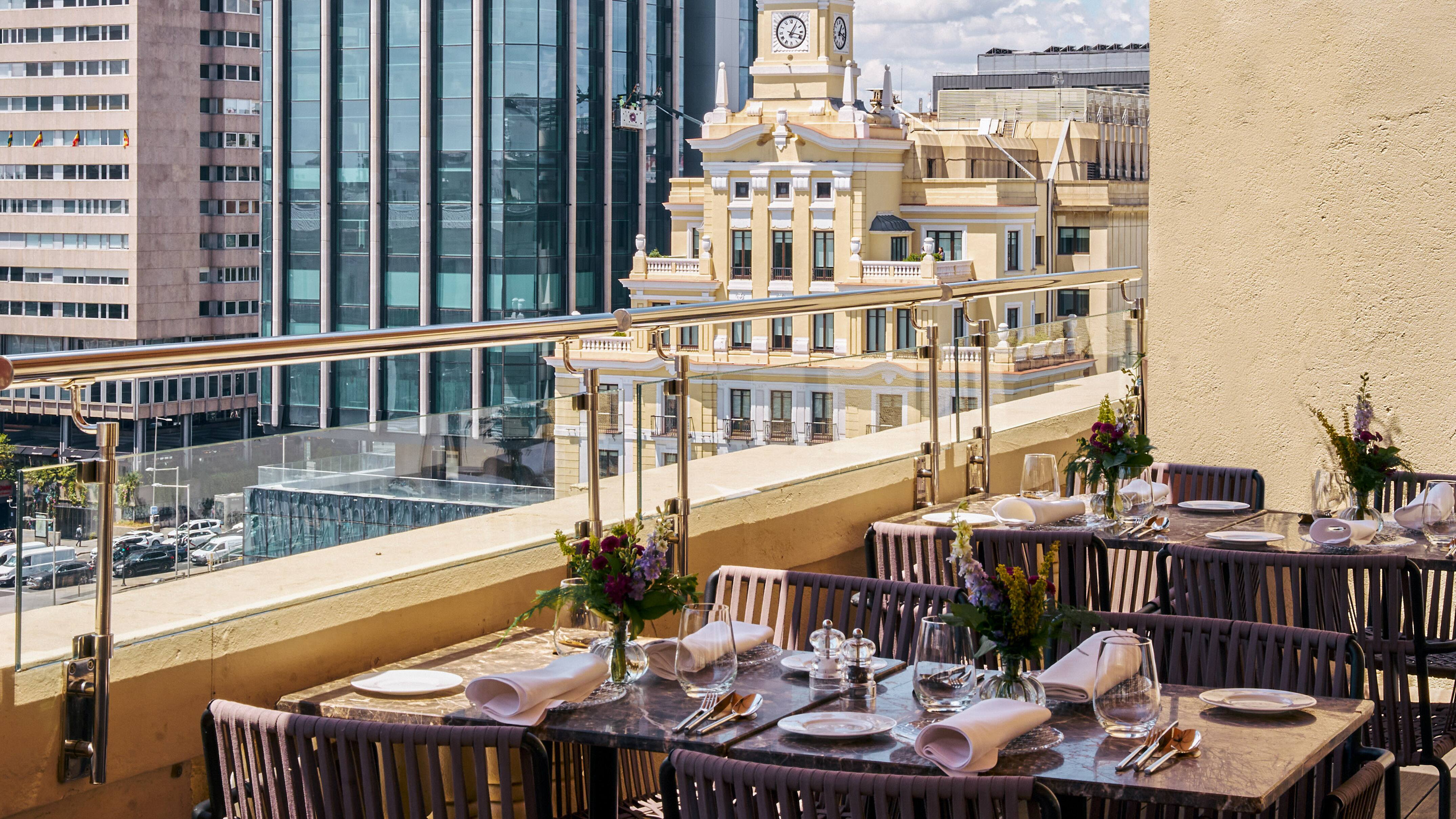 An outdoor terrace with marble-topped tables set with white napkins, glassware and small flower vases behind a glass balustrade, overlooking a yellow clock-towered building and glass office blocks.