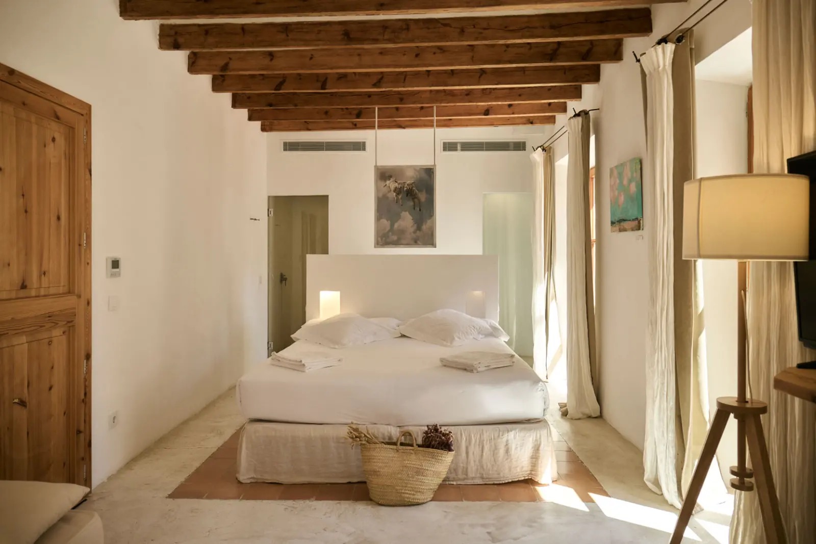 Guest room with an exposed wood-beam ceiling and a white bed dressed with linen and a woven basket