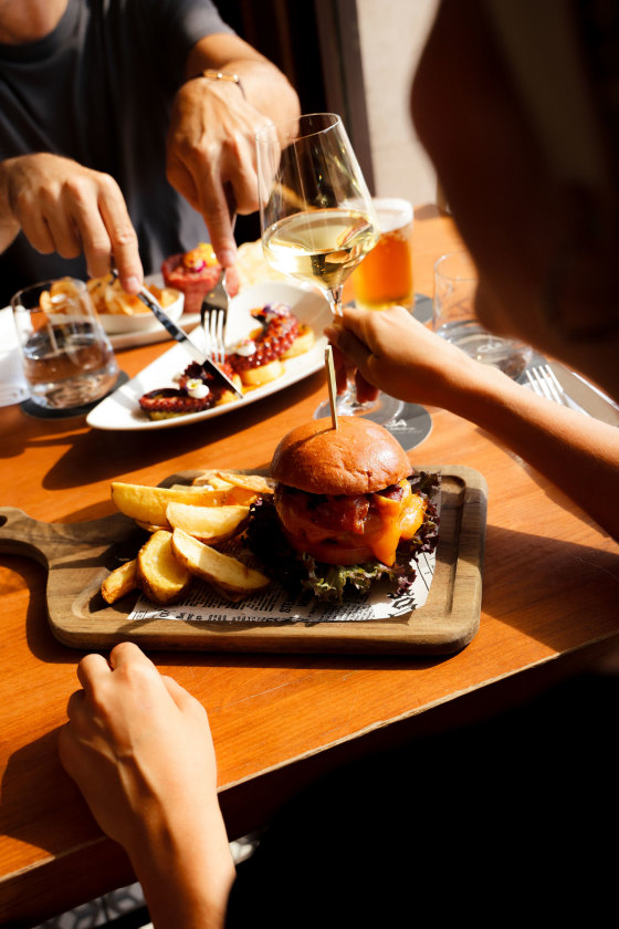 A burger and fries served on a wooden board at the hotel restaurant, alongside a plate of octopus and a glass of white wine