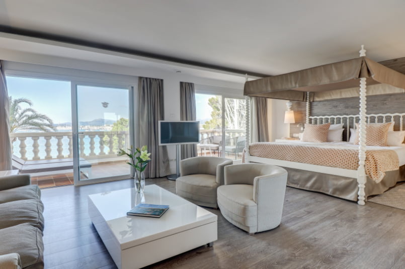 Suite bedroom with a white four-poster bed, a lounge area and a balcony overlooking the bay