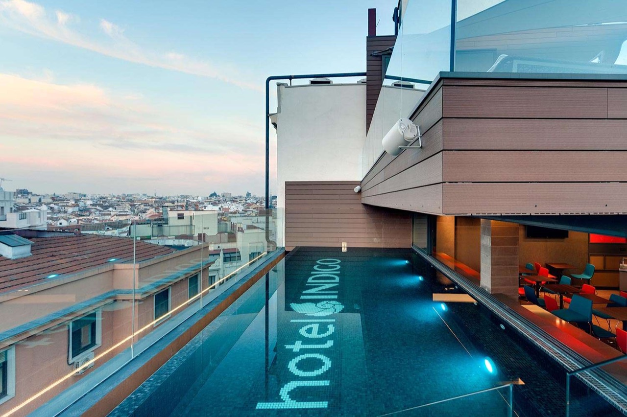 Rooftop infinity pool with 'Hotel Indigo' tiled into the floor, glass railing and Madrid rooftops and the Torre de Madrid in the background.