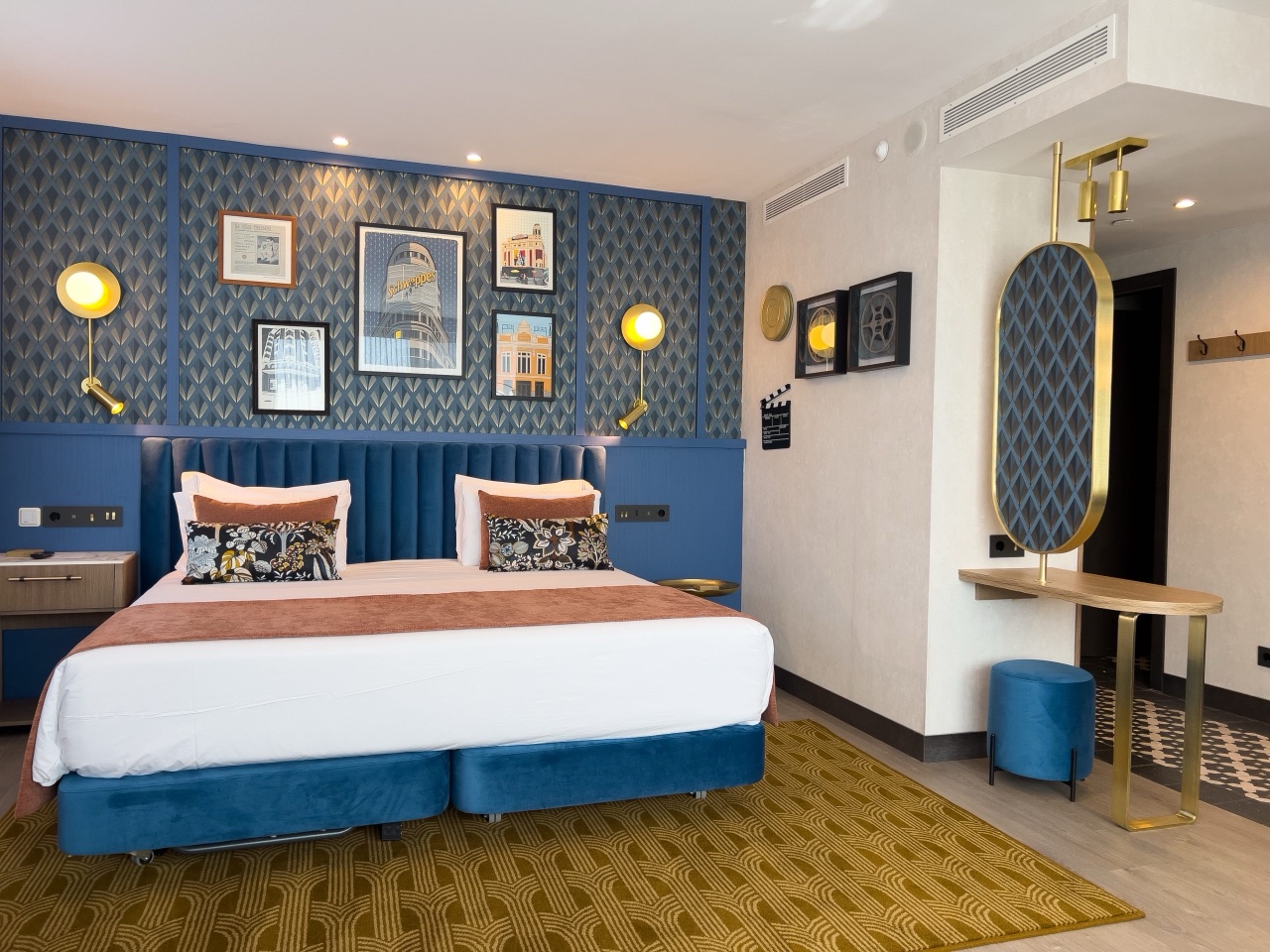Hotel room with a blue upholstered headboard, framed Madrid-themed art prints and an art-deco patterned rug.