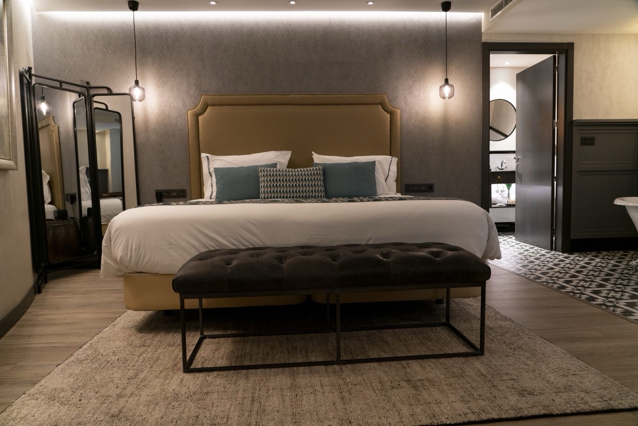 Hotel room at night with a mustard-yellow upholstered headboard, hanging pendant lights and a leather bench at the foot of the bed, bathroom visible through an open door.